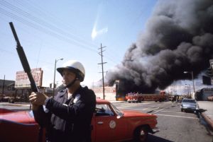 A police officer stands guard to secure a perimeter in the Watts neighborhood as members of the Los Angeles Fire Department battle a fire that engulfed a local business in the wake of ongoing riots, Los Angeles, California, mid-August. 1965. (Photo by Lawrence Schiller/Polaris Communications/Getty Images)