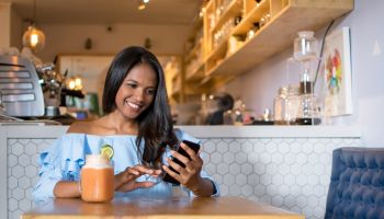 Black woman at a cafeteria drinking a smoothie and looking at her smartphone