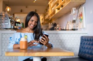 Black woman at a cafeteria drinking a smoothie and looking at her smartphone