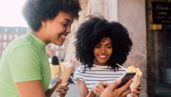 Two African - American friends eating ice cream and using a smartphone on the street