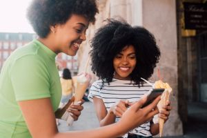 Two African - American friends eating ice cream and using a smartphone on the street