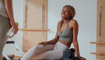 Beautiful African smiling sportswoman sitting on pilates reformer and chatting with her girlfriend.