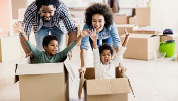 Happy black parents having fun while pushing their kids in cardboard boxes at new apartment.