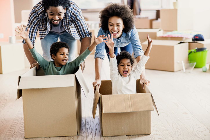 Happy black parents having fun while pushing their kids in cardboard boxes at new apartment.