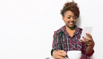 Black young woman sitting down on floor checking her social media enjoying coffee
