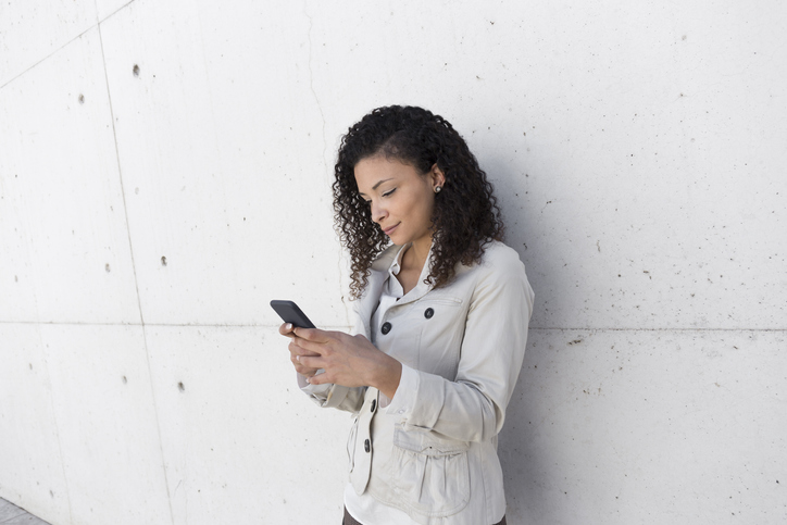 Stylish businesswoman using smart phone while standing against wall