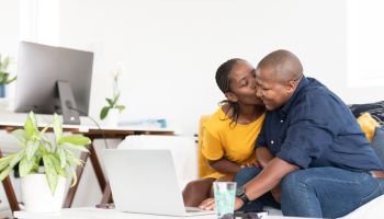 Woman kisses her partner sitting on sofa using laptop together
