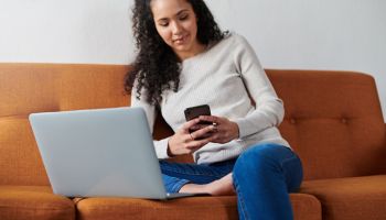 Shot of a young female sitting on the couch, using her laptop and phone at home