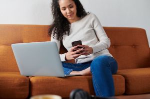 Shot of a young female sitting on the couch, using her laptop and phone at home