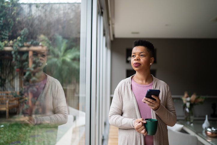 Woman contemplating holding a mug and a smartphone at home