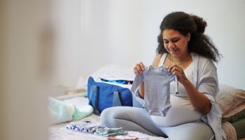 Mother, planning and pregnant with woman with baby clothes for birth suitcase checklist for family, healthcare and love. Pregnancy, maternity and packing bag of newborn clothing in bedroom at home