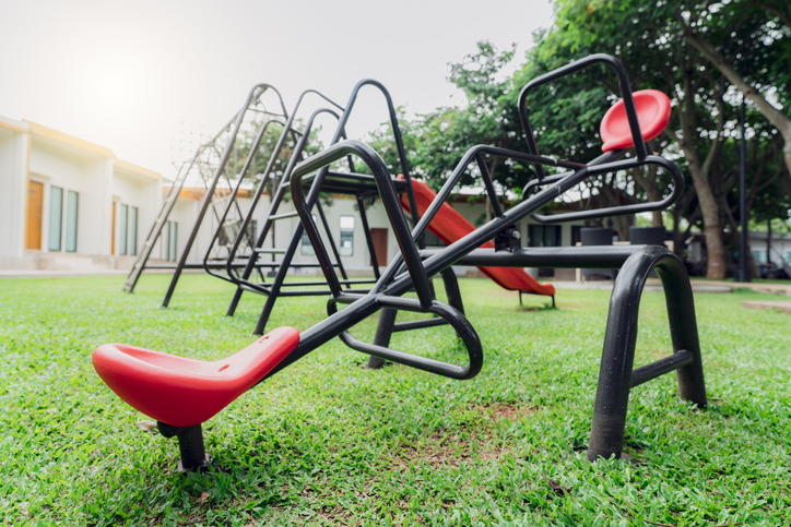 Red seesaw in the playground. Playground equipment for children to play. Plastic seesaw or teeter-totter, swing, and slide at outdoor playground with green grass ground. Outdoor kids toy at park.