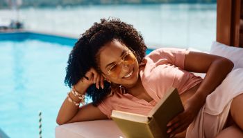 Happy black woman reading book while relaxing on deck chair by the pool.