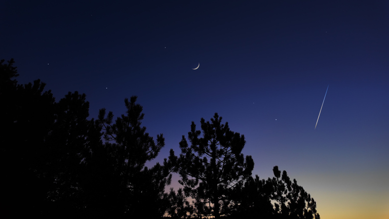 Countryside silhouettes under the stars, meteor trail and crescent Moon.
