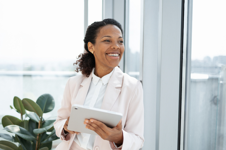 Confident professional woman smiling while holding a tablet near a window