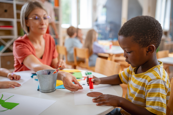 Preschool teacher assisting diverse children with arts and crafts using scissors, glue, and colorful paper in a bright, cheerful classroom