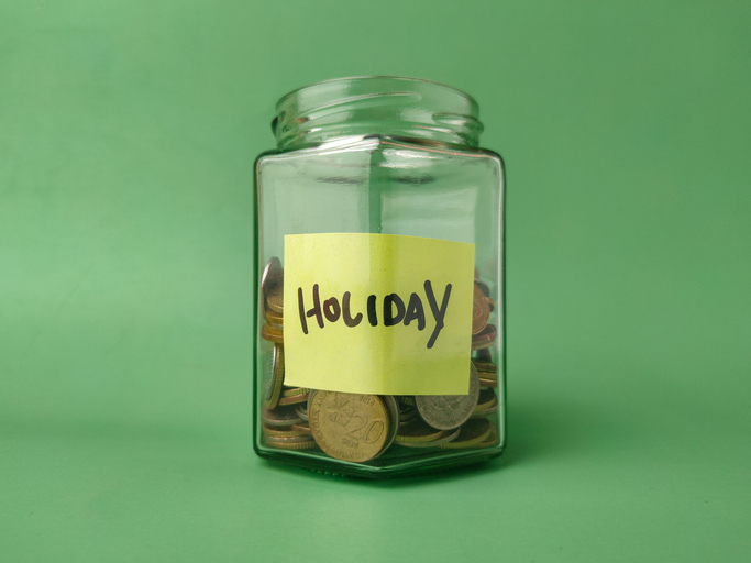 The image shows a glass jar filled with coins and a yellow sticky note on it with the word Holiday written in black marker