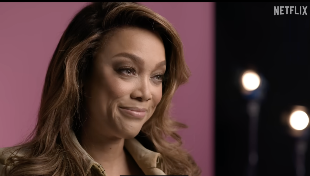 A woman with long brown hair and a warm smile, wearing a black jacket, against a pink background with the Netflix logo visible.