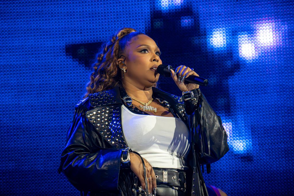 A woman with curly hair singing into a microphone on stage, wearing a black leather jacket with studs and a white top.