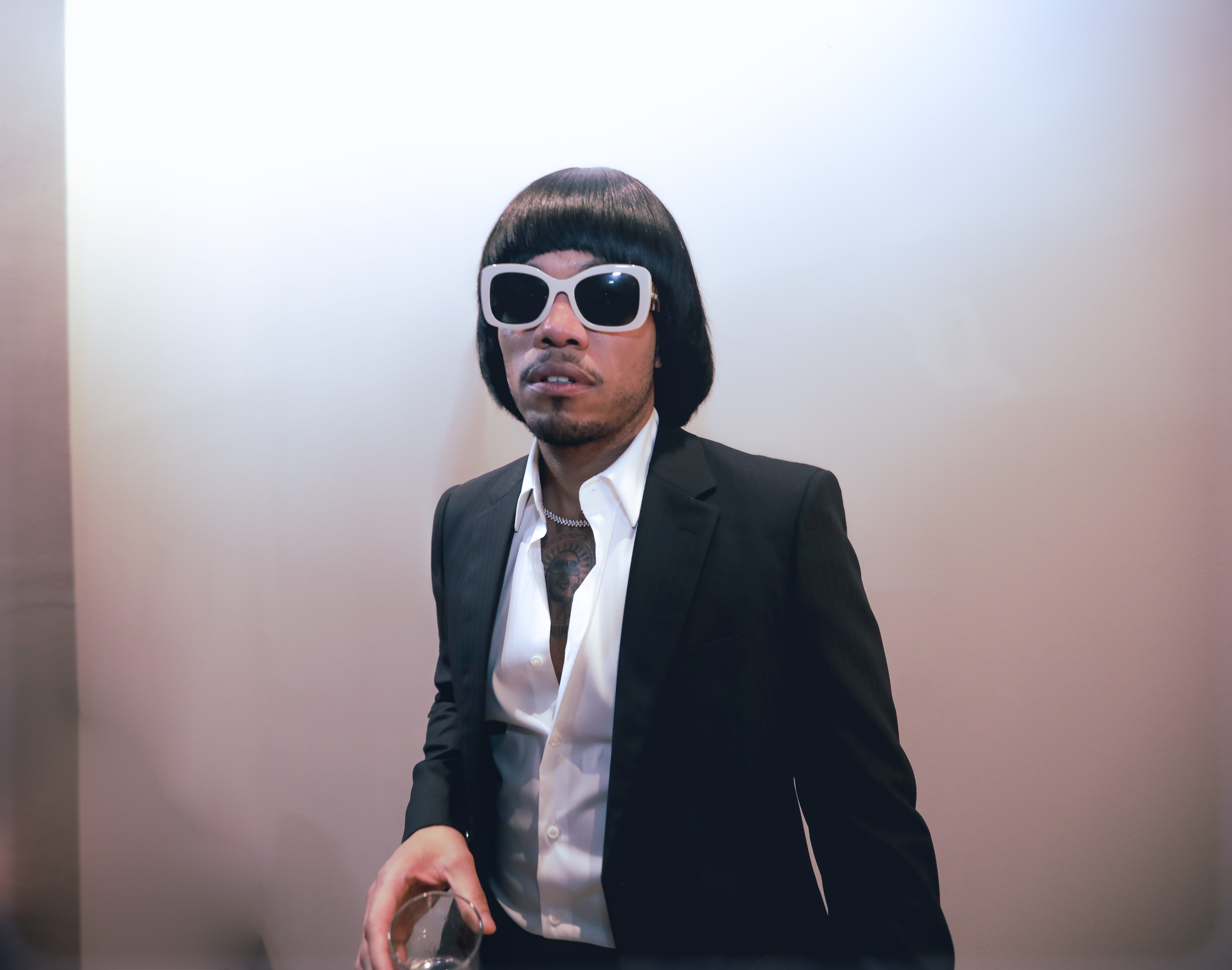 A person wearing a black suit, white shirt, and sunglasses stands in a room with a blurred background.