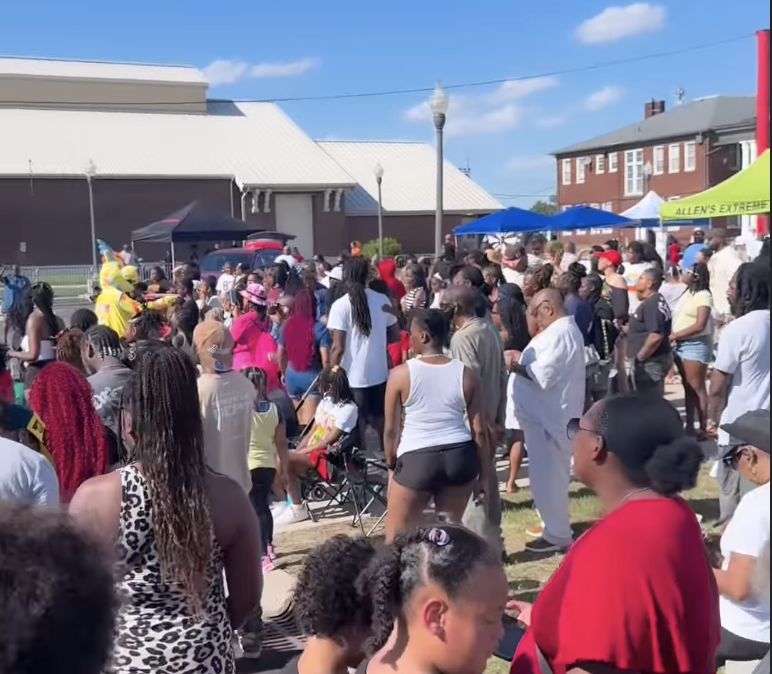 A crowded outdoor event with people of diverse backgrounds gathered together, some wearing colorful clothing and accessories.