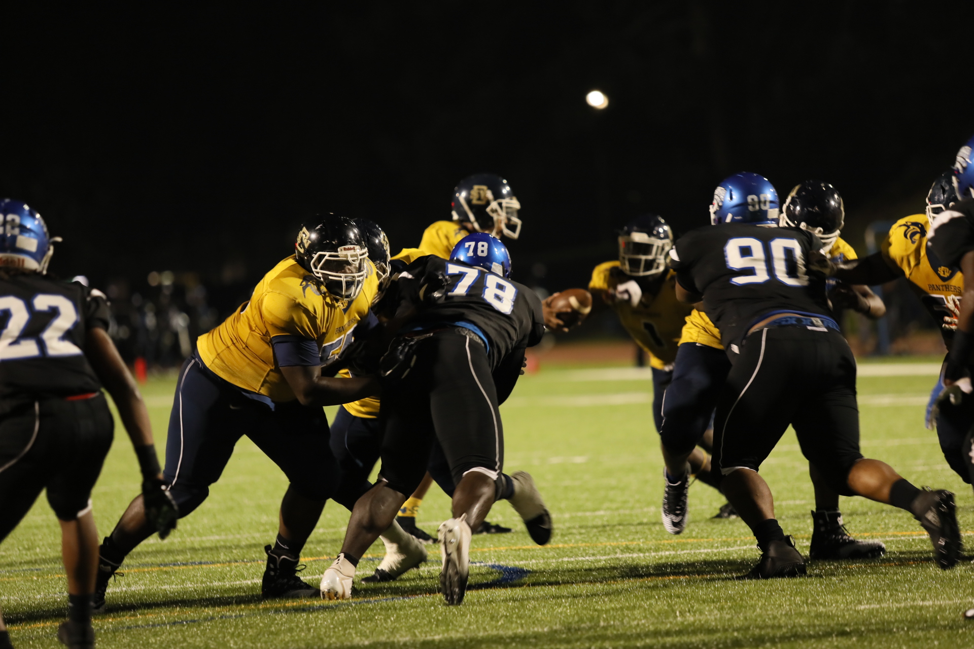 Football players in a night game, with players in yellow and blue uniforms engaged in a play on the field.
