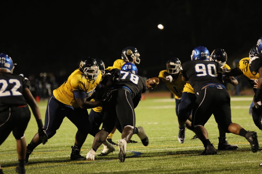 Football players in a night game, with players in yellow and blue uniforms engaged in a play on the field.