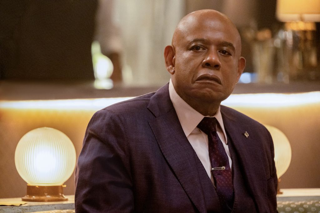 A serious-looking bald Black man in a dark suit and tie, sitting in what appears to be a hotel or office setting.