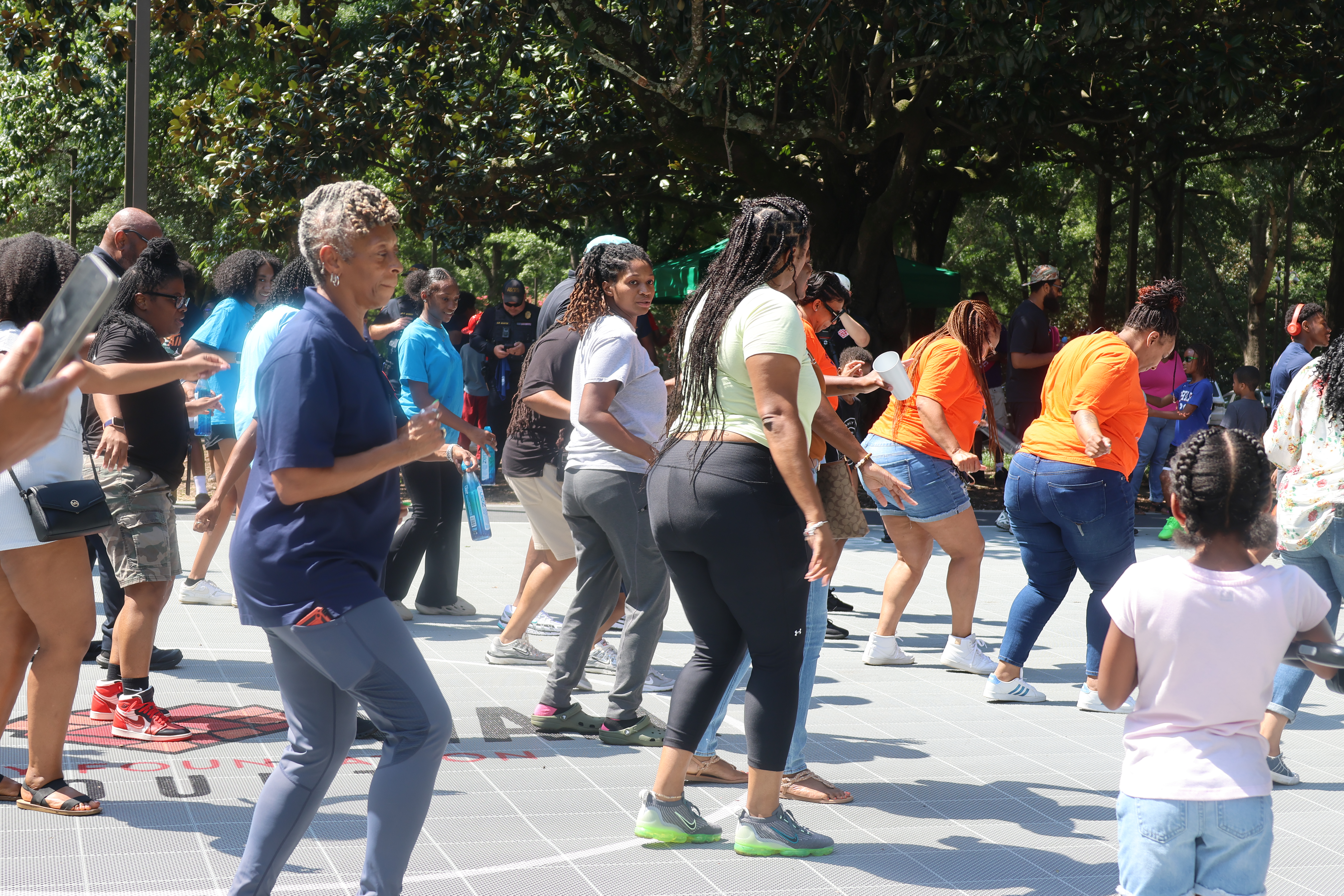 A group of people dancing outdoors in a park-like setting, with trees and greenery in the background.