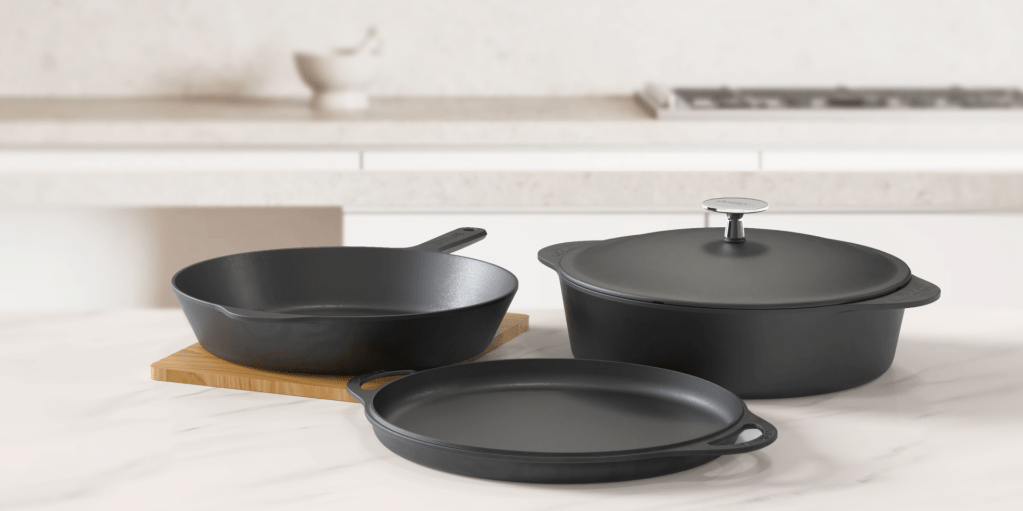 Black cast iron cookware including a skillet, Dutch oven, and baking dish on a wooden board against a white background.