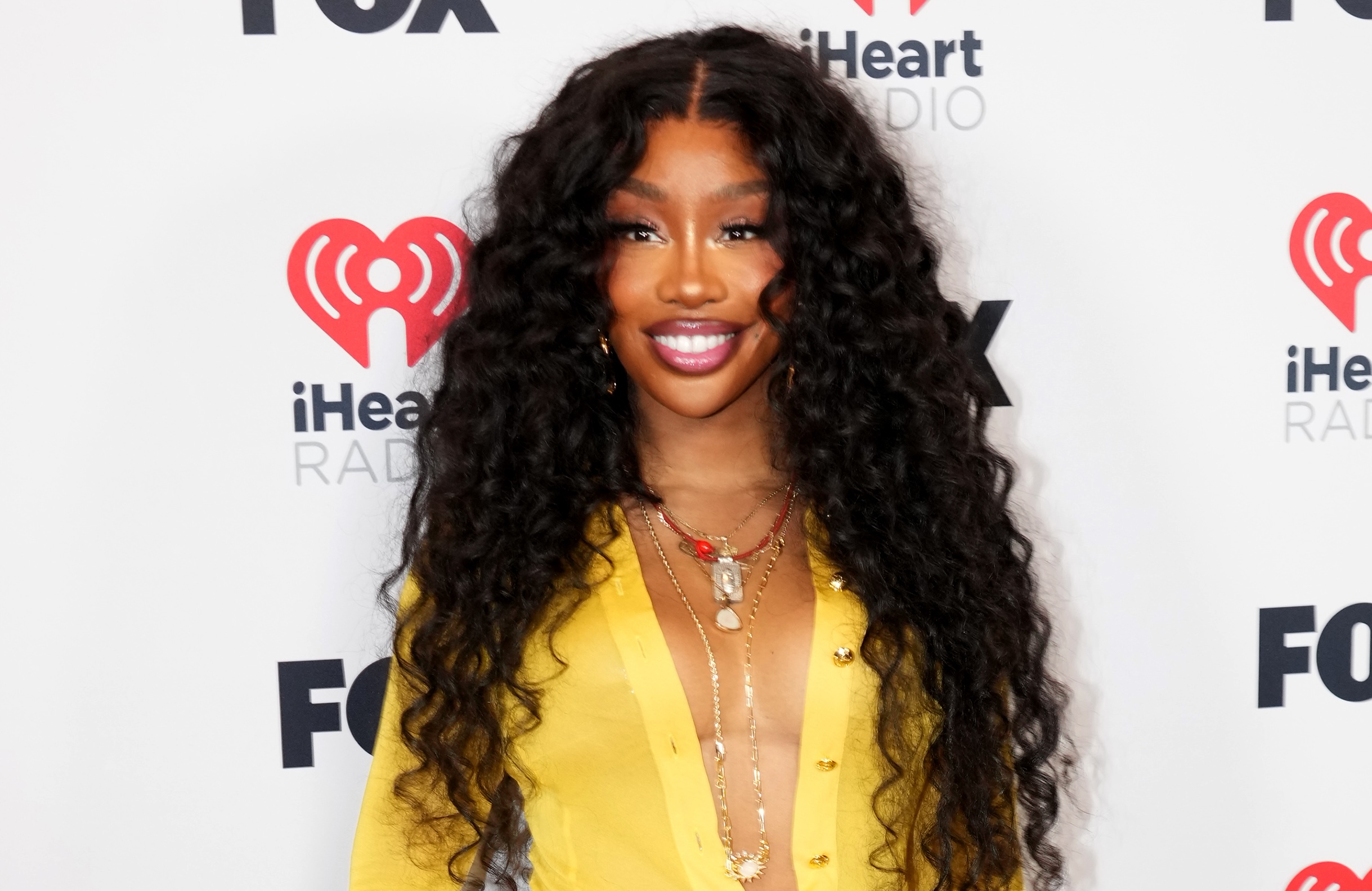 A woman with long, curly black hair wearing a yellow jacket and necklaces posing in front of a backdrop with "iHeart Radio" and "FOX" logos.