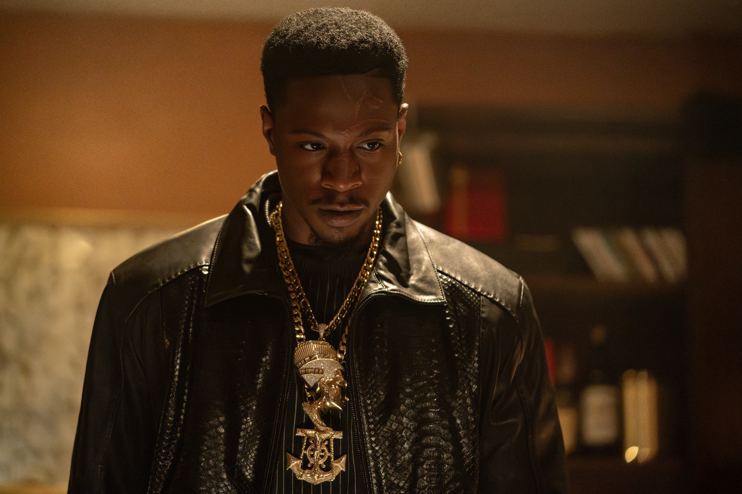 A close-up portrait of a serious-looking Black man wearing a black leather jacket, gold chains, and a gold pendant.
