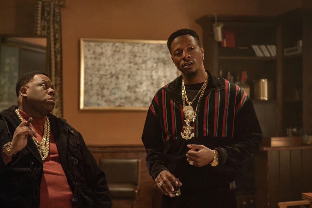 Two African American men wearing gold jewelry and stylish clothing standing in a room with shelves and artwork.