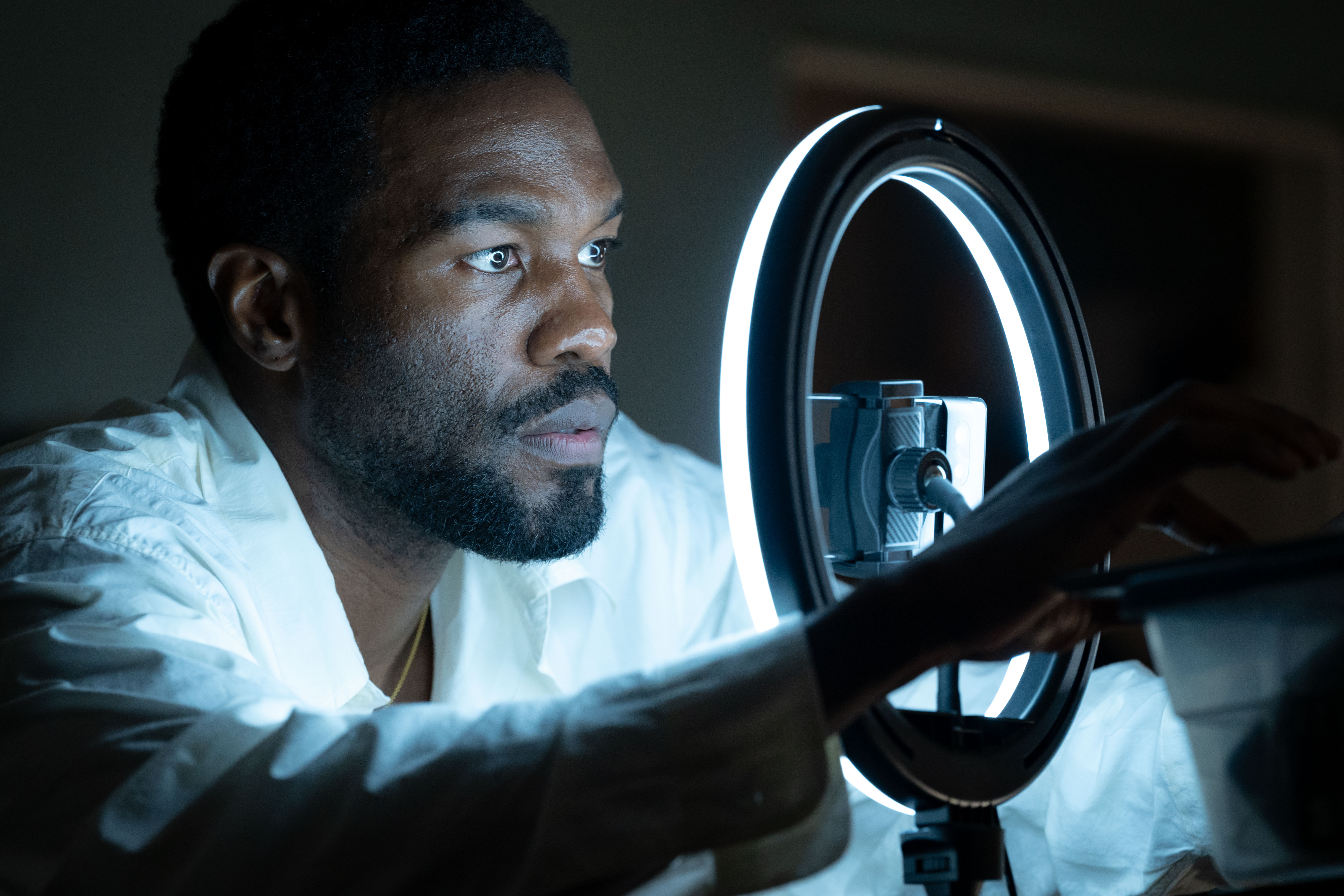A man with a beard and serious expression looking into a mirror in a dimly lit room.