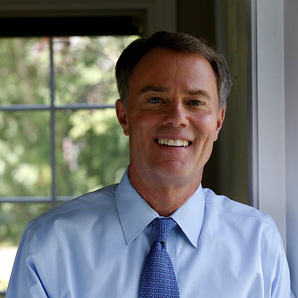A smiling middle-aged man in a blue shirt and tie standing in front of a window.