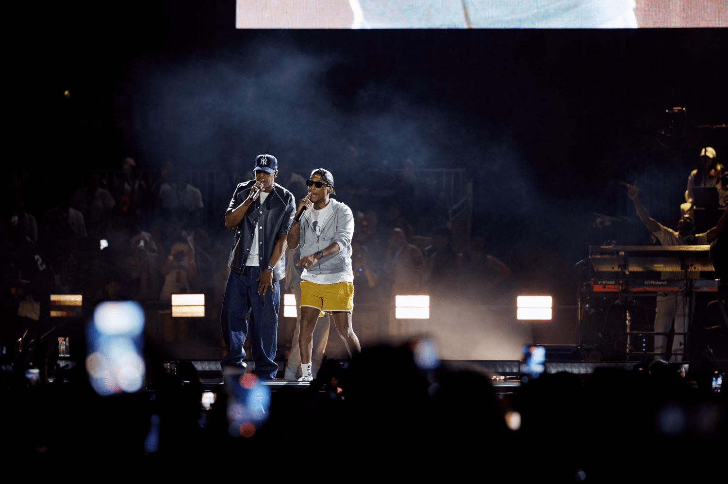 Two performers on stage in a smoky, dimly lit venue, one wearing a Yankees cap and the other in yellow shorts.