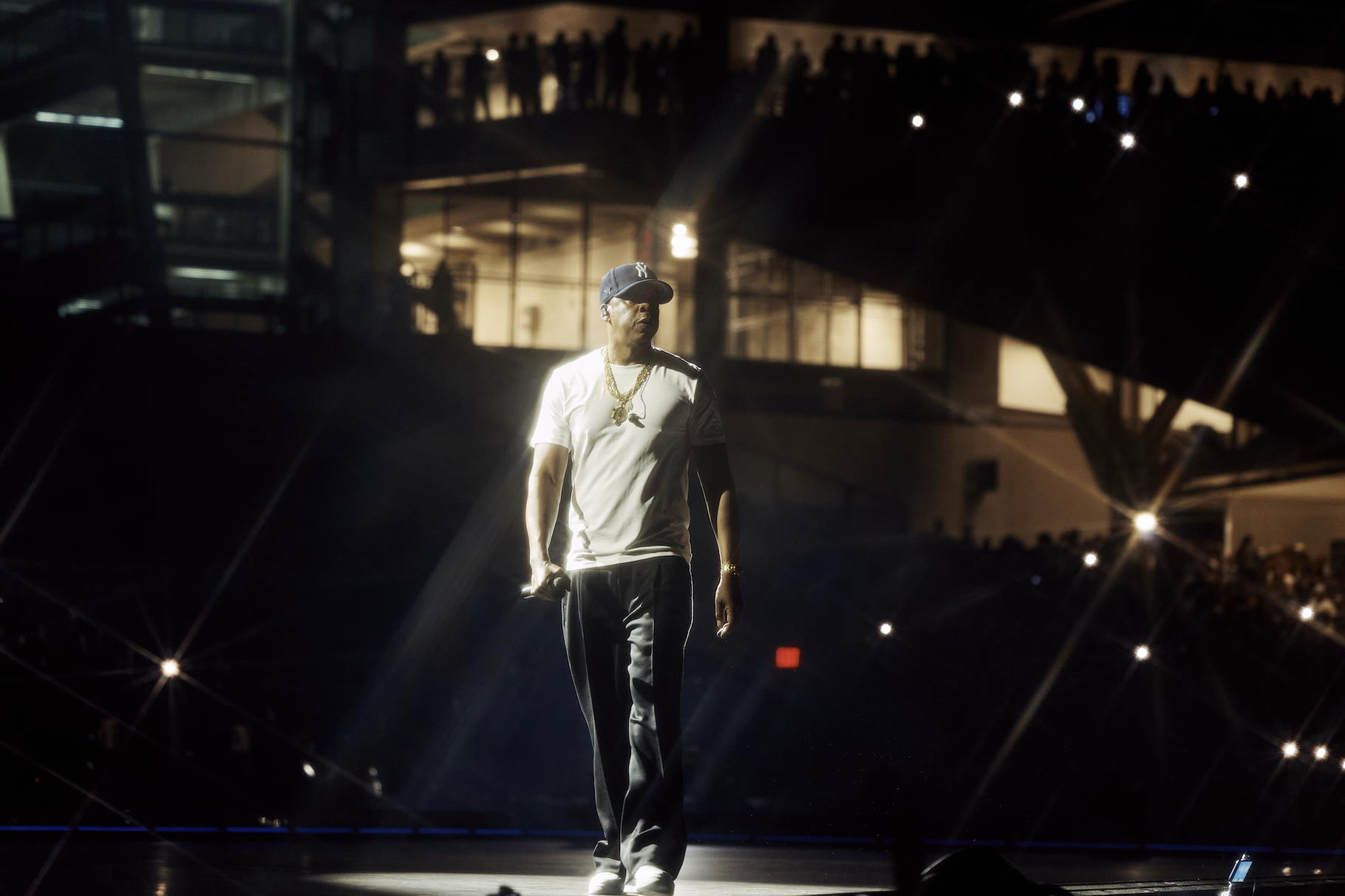 A person wearing a white shirt, baseball cap, and jeans standing on a stage in a dimly lit venue.