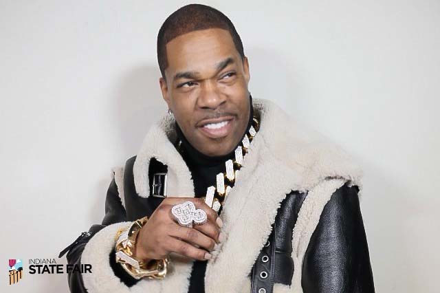 A smiling man wearing a fur-trimmed jacket and gold jewelry, holding a microphone.