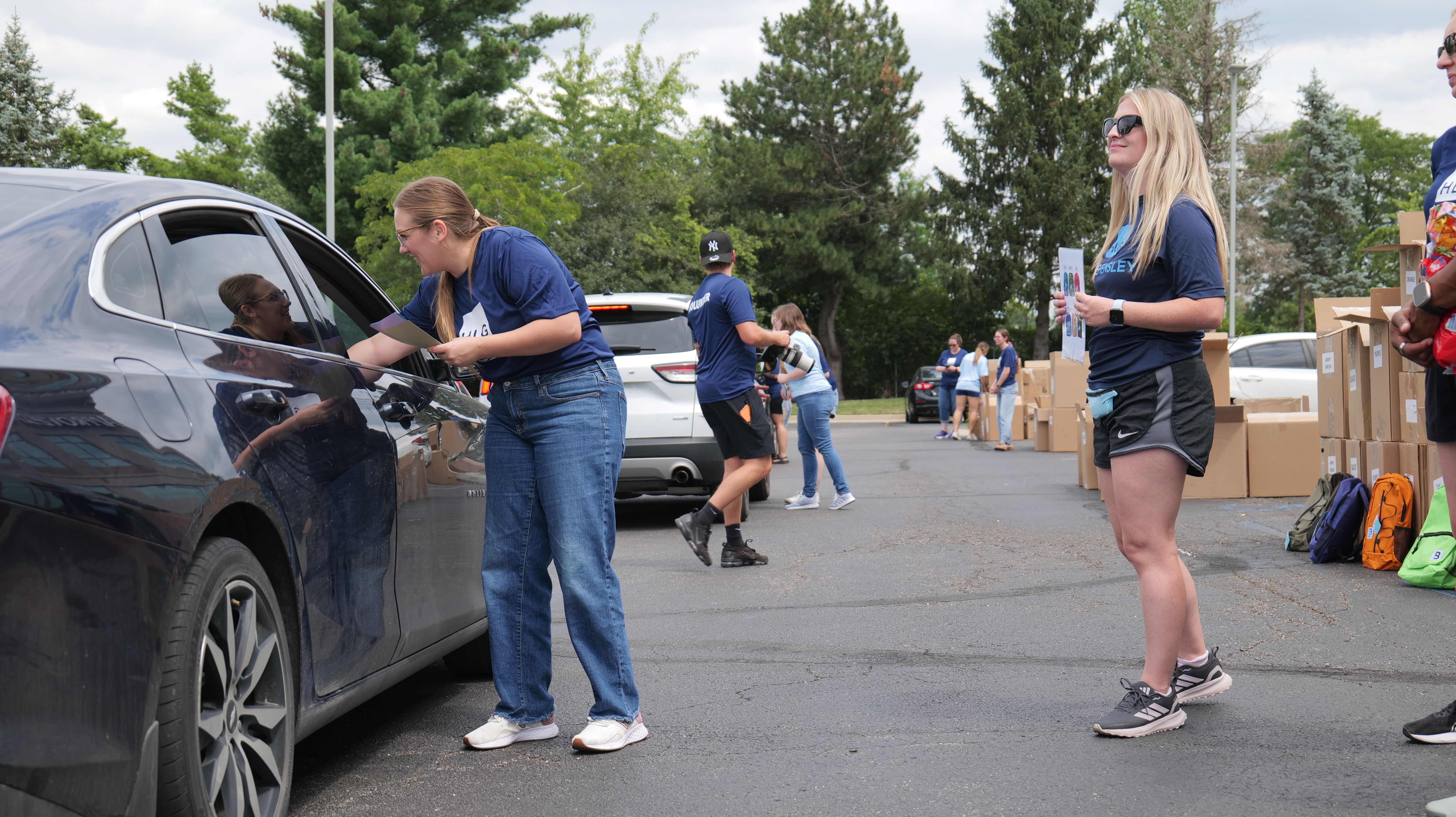 A group of people unloading boxes from a car in a parking lot, with a large vehicle in the background.