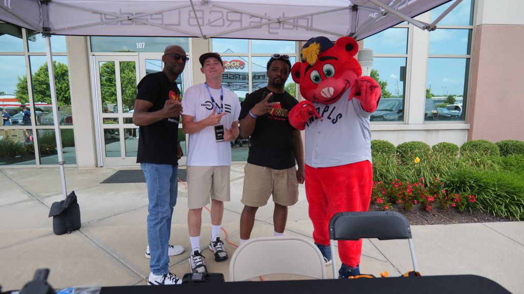 Four people, including a person in a red mascot costume, posing for a photo in front of a building with "107" visible.