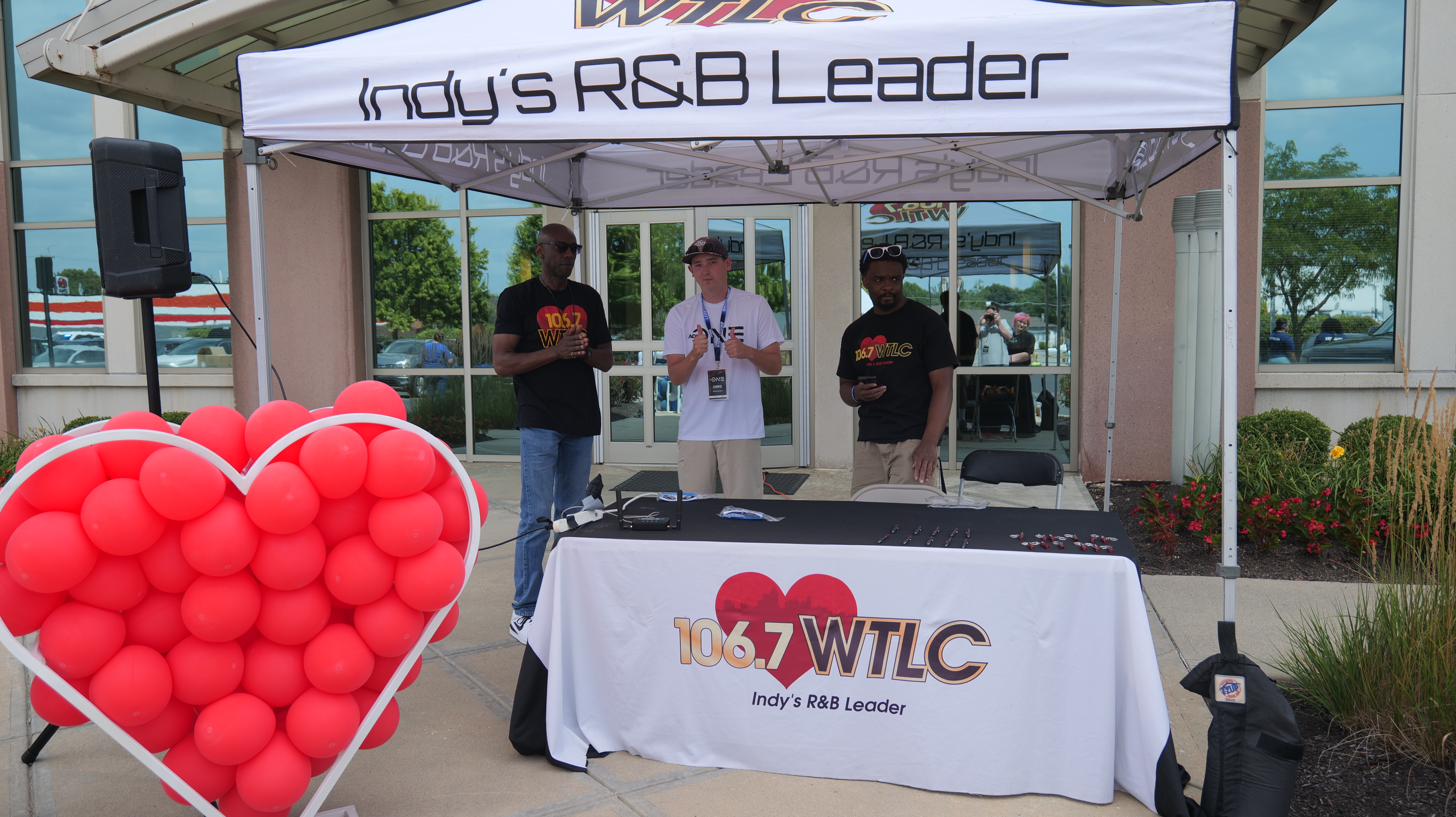 A tent with the banner "Indy's R&B Leader" and the 106.7 WTLC logo, with three people standing behind a table with red heart-shaped balloons.
