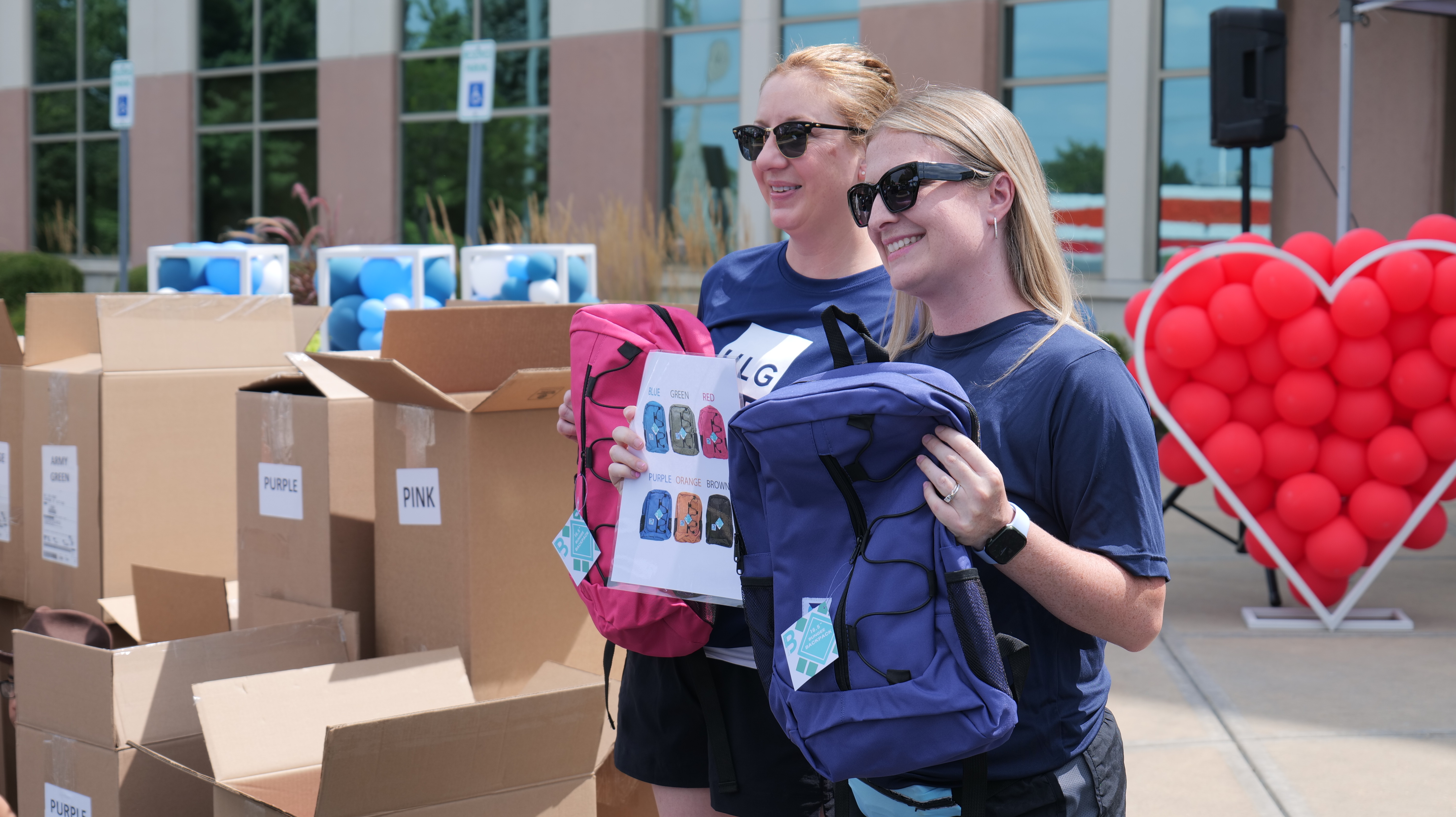 Two young women wearing matching blue shirts and backpacks, smiling and standing in front of boxes and red heart-shaped balloons.