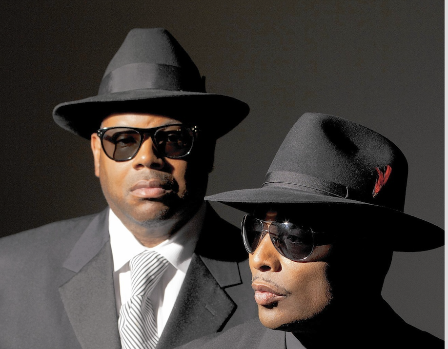 Two men wearing hats and sunglasses, one in a suit and the other in a wide-brimmed hat, standing together against a dark background.