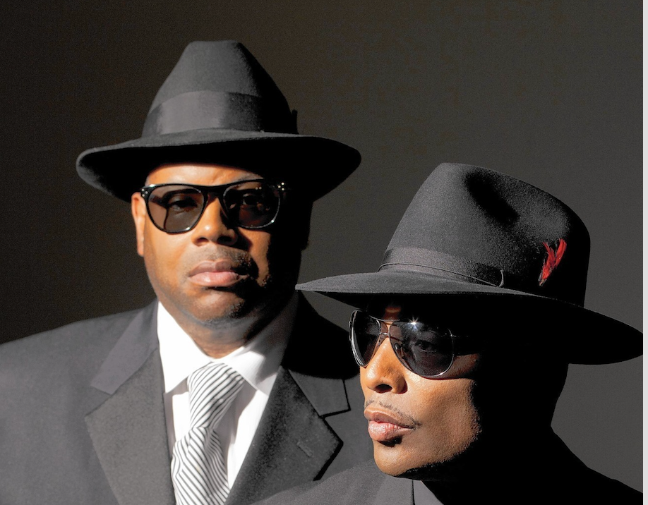 Two men wearing hats and sunglasses, one in a suit and the other in a wide-brimmed hat, standing together against a dark background.