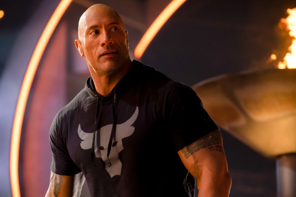 A muscular man in a black shirt with a logo stands in a dramatic setting with fiery lights.