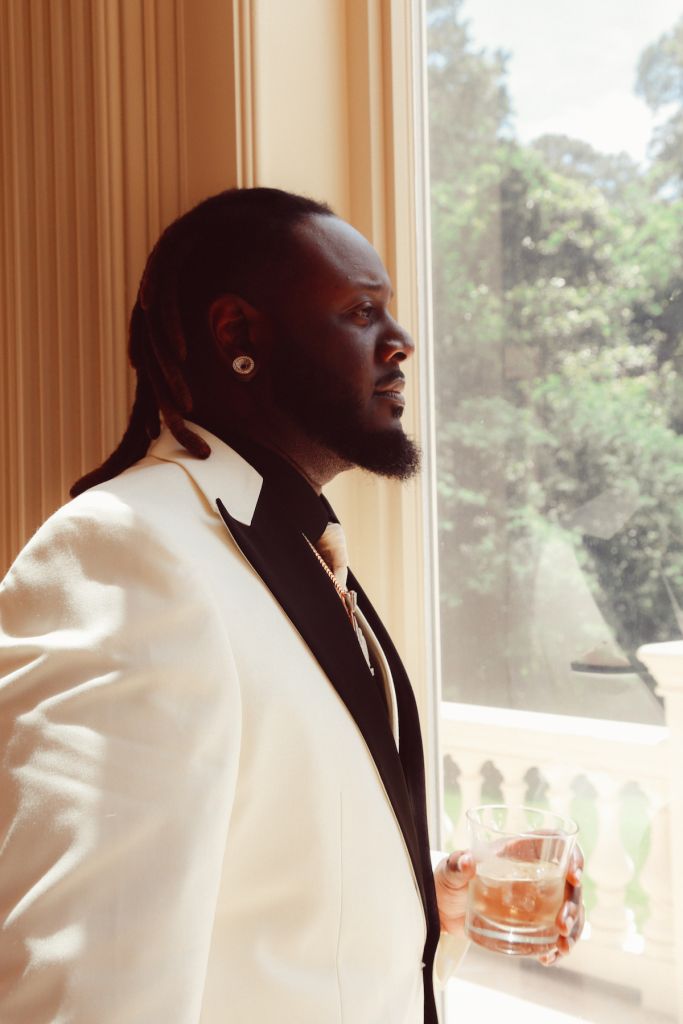 A man in a white suit standing by a window, holding a glass of liquor and looking out at the trees outside.