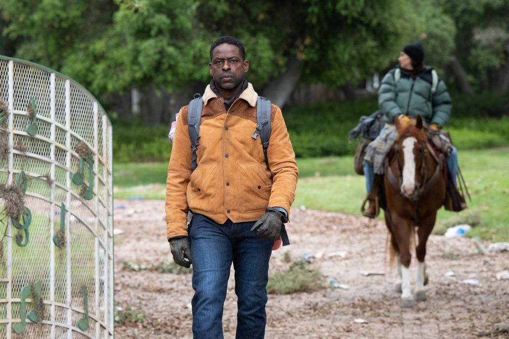 A man wearing a yellow jacket and backpack walking on a dirt path, with a person riding a horse in the background.